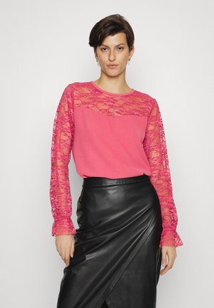 Blusa rosa com mangas longas de renda, apresentando um decote redondo e tecido texturizado. Combinada com uma saia envolvente preta de couro.
