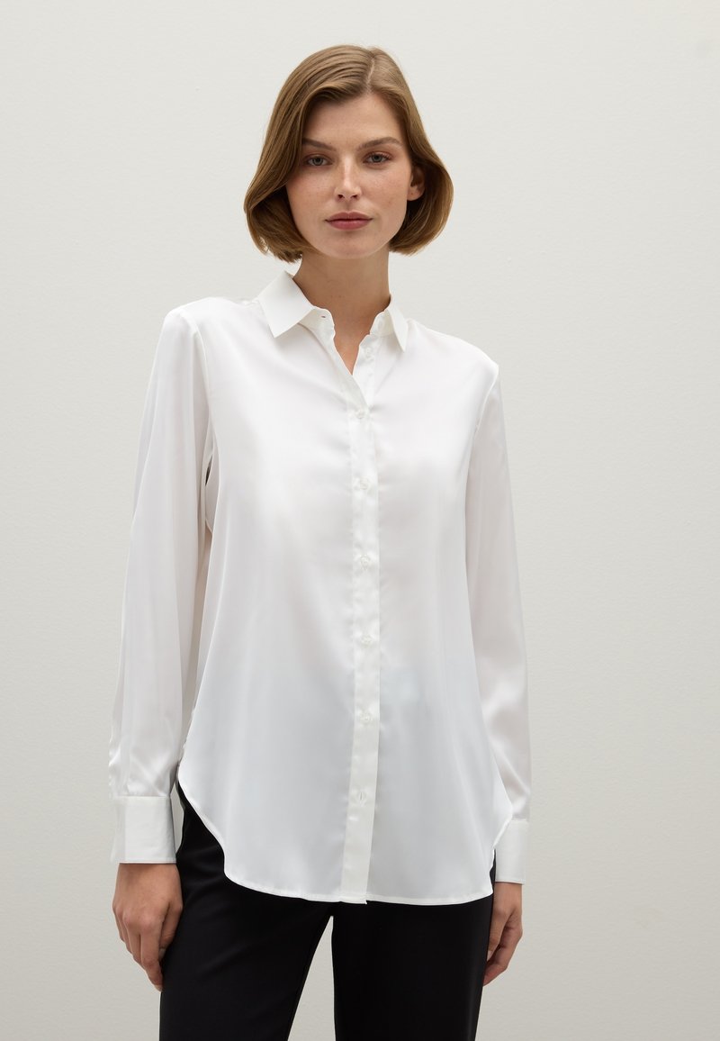 Camisa blanca de manga larga con cuello estándar, frente de botones, textura suave similar a la seda, dobladillo curvado y puños ajustados. Llevada con pantalones negros.
