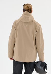 Beige wasserdichte Jacke mit Kapuze, langen Ärmeln und lockerer Passform. Die Person hält einen schwarzen Helm in der linken Hand.
