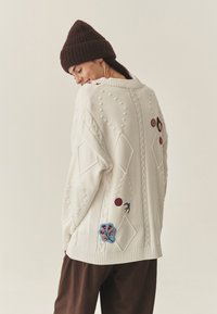 Przesadnie duży biały sweter z dzianiny, ozdobiony wzorem w warkocze, teksturowanymi kulkami oraz kolorową, kwiatową haftowaną dekoracją. Uzupełniony brązową czapką typu beanie.