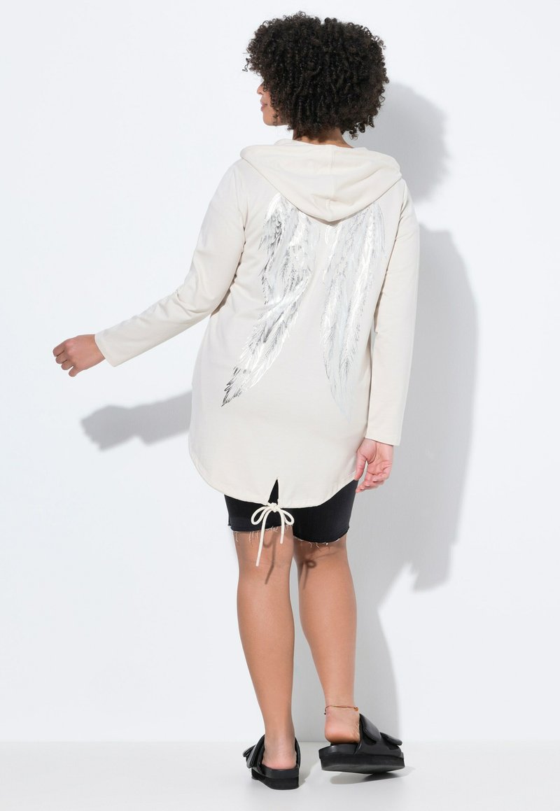 Hoodie lungo beige con un design di ali argentate sul retro, dotato di orlo con coulisse e spacco laterale, abbinato a pantaloncini di jeans scuri.