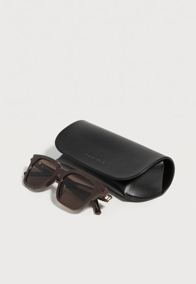 Lunettes de soleil carrées brun foncé avec des verres foncés à côté d’un étui de protection en cuir noir fermé sur une surface blanche.