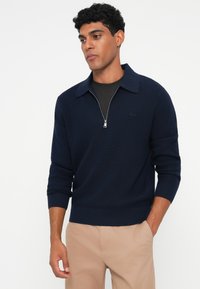 Pulover cu guler tip fermoar în nuanță de bleumarin, cu textură cu nervuri și guler structurat, având un logo subtil și mâneci lungi, asortat cu pantaloni bej deschis.