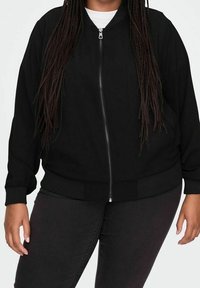 Giacca bomber nera con zip frontale, polsini e orlo a costine. Presenta una vestibilità ampia e un tessuto liscio, offrendo un design minimalista.