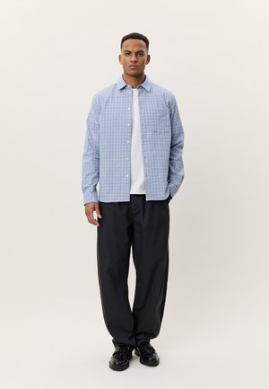 Muž stojící, má na sobě světle modrou kostkovanou košili přes bílé tričko, volné černé kalhoty a černé boty na jednoduchém pozadí.