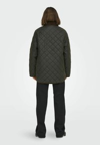 Giacca trapuntata verde scuro con colletto alto, caratterizzata da una forma rettangolare, vestibilità ampia e bordi rifiniti, indossata sopra pantaloni neri.