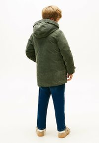 Grøn kordjakke med hætte, der har en lodret ribbet tekstur, parret med blå denimbukser og beige sko.