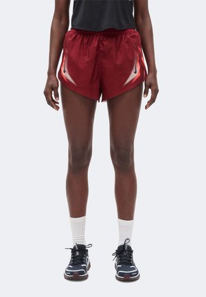 Ben på en person iført burgundy sportsshorts med hvidt og sort design, sorte sneakers med hvide såler og hvide crew-sokker.
