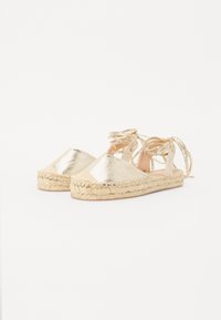 Kulta metalliset espadrillokset, joissa on punotut juutti-pohjat, mantelin muotoinen kärki ja ristikkäinen nauhakiristys. Litteä, avonainen takatyyli.