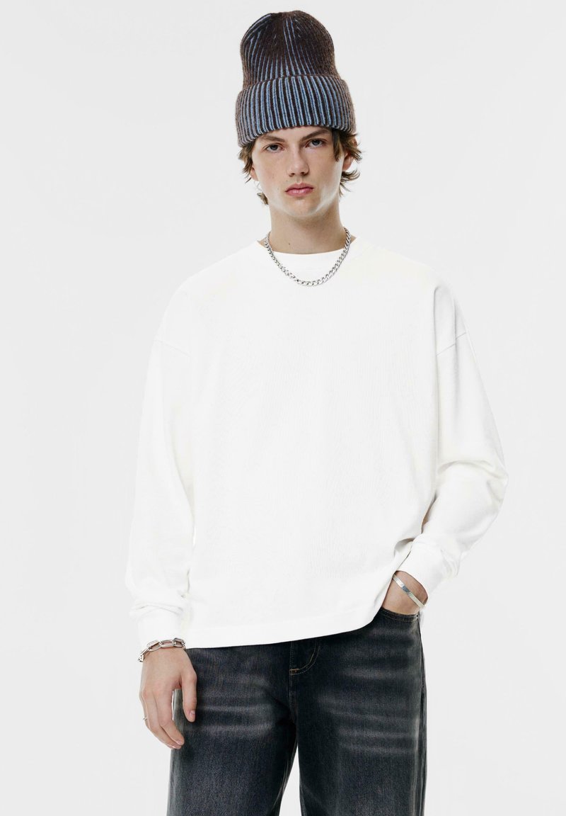 Camicia bianca a maniche lunghe con texture a coste, indossata con jeans di denim scuro. Un berretto a maglia grigio a righe e una collana in argento completano il look.