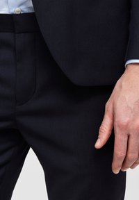 Marina kostymbyxor i strukturerad tyg, med rak skärning, platt framsida och sidofickor. Närbild av hand som vilar på tyget.