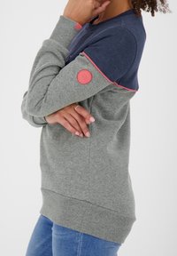Gray sweatshirt with a navy upper section, pink accents, and a round logo patch on the sleeve. Soft material, relaxed fit, ribbed cuffs.
