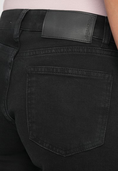 Nærbillede af en person i sorte denim jeans med en baglomme og et sort læder mærke på taljen.