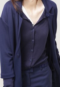 Marinblå långärmad cardigan över en matchande knäppblus. Texturerad tyg med en mjuk yta. Outfiten är lager-på-lager och avslappnad.