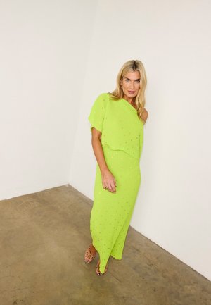 Donna con lunghi capelli biondi che indossa un vestito verde neon monospalla con pois dorati e sandali in pelle di serpente, in piedi su un pavimento in cemento.