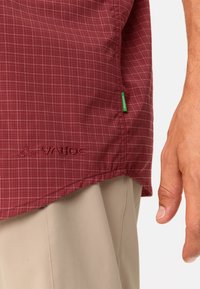 Camisa de cuadros rojos con el logo bordado "Vaude", que cuenta con una etiqueta lateral en verde. La tela parece texturizada y ligera.