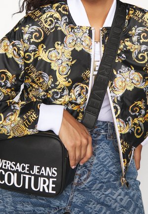 Personne portant une veste noire et or à motifs ornés, un jean bleu, et tenant un sac bandoulière noir Versace Jeans Couture.