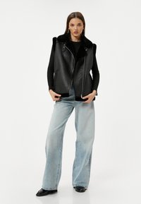 Schwarze Lederweste mit Kunstfellkragen, Reißverschluss vorne und seitlichen Taschen, kombiniert mit hellblauen weiten Jeans und schwarzen Schuhen.