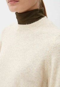 Frau trägt einen mehrlagigen cremefarbenen Strickpullover über einem dunkelolivgrünen Rollkragenpullover, der den Hals und das untere Gesicht zeigt.