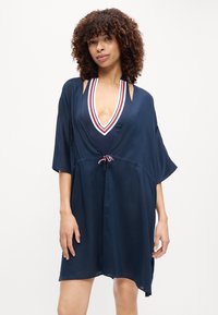 Γυναίκα με σγουρά μαλλιά που φοράει μαγιό σε σκούρο μπλε με V-λαιμόκοψη και αντίστοιχο χαλαρό κάλυμμα με κορδονάκι στη μέση.