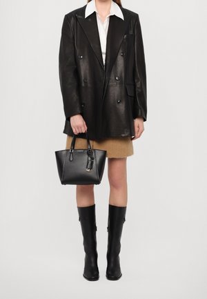 Femme portant un blazer en cuir noir à double boutonnage, une chemise blanche, une jupe marron, des bottes noires montant jusqu'aux genoux, tenant un sac à main en cuir noir.