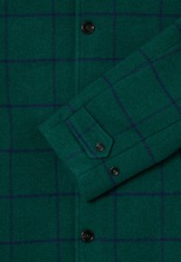 Grønn ullskjortejakke med blå rutemønster, med knappede mansjetter og svarte knapper. Myk tekstur, lett børstet overflate.