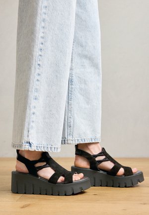 Füße, die schwarze Plateausandalen mit dicken Sohlen und Riemen tragen, kombiniert mit hellblauen Jeans mit weitem Bein, stehend auf Holzboden.