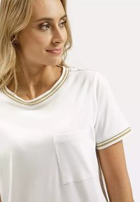 Biała bawełniana koszulka z krótkim rękawem, z okrągłym dekoltem i kieszenią z przodu. Uzupełniona złotymi paskami wykończenia przy kołnierzyku i rękawach.