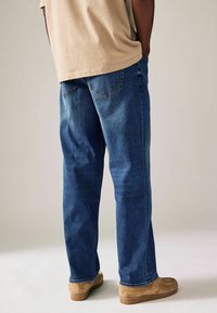 Jeans de denim azul con corte relajado, perneras rectas y bolsillos traseros. Combinados con una camisa beige clara y calzado color marrón claro.
