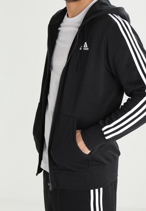 Homme portant un survêtement Adidas noir à rayures blanches, T-shirt blanc, mains dans les poches de la capuche, debout devant un fond clair uni.