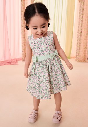 Joven niña sonriendo y mirando hacia abajo, vestida con un vestido floral con lazos y sandalias rosas en una habitación de colores pastel.