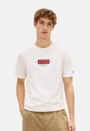 Joven con cabello rizado que lleva una camiseta blanca de Tommy Hilfiger con logo rojo y pantalones beige, de pie frente a un fondo liso.