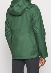 Giacca impermeabile verde con cappuccio, caratterizzata da una texture liscia e un design aderente. Il retro presenta dettagli delle cuciture che mettono in risalto la sua forma.