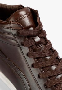 Zapatilla alta de cuero marrón con superficie texturizada, cordones marrones y detalles de costura. Presenta un logotipo grabado en el talón.