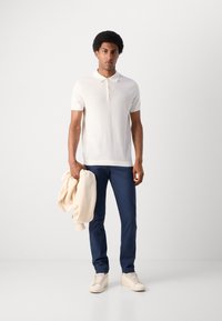 Polo shirt blanc texturé à manches courtes, associé à un pantalon bleu foncé. La personne tient un pull crème, portant des baskets de couleur claire.