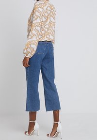 Blouse à manches longues à motifs en chaînes blanches et dorées, associée à un jean cropped bleu. Le modèle porte des sandales à talons blanches avec des brides cheville.