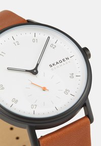 Rund vit urtavla med svarta tim- och minutvisare, orange sekundvisare, svart metallkapsel och brun läderrem med synlig textur.