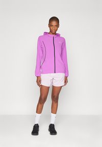 Lätt rosa zip-up huvtröja med svarta detaljer och långa ärmar, i kombination med ljusrosa shorts och svarta träningsskor.