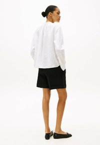 Camisa de lino blanca de manga larga, vista trasera, combinada con pantalones cortos negros a medida. Zapatos planos negros con un lazo decorativo. Fondo liso.