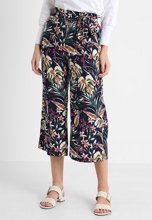 Kobieta ubrana w granatowe, szerokie spodnie o długości do pół łydki z kolorowym kwiatowym wzorem oraz białe sandały z klamrami.