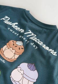 Mørk teal T-shirt med legende dessertillustrationer og et hvidt logo, der står "Pusheen Macarons sweet every day." Blødt, bomuldsstof.