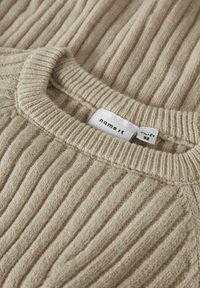 Pull en tricot côtelé beige avec un col rond. La texture du tissu est douce avec des lignes verticales. Étiquette visible avec le nom de la marque "name it" et la taille.