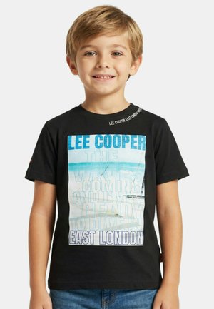 Jeune garçon souriant aux cheveux blonds portant un T-shirt noir Lee Cooper avec un motif graphique bleu et blanc sur le thème des vagues et de l'Est de Londres.
