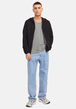 Giovane uomo in piedi, indossa una felpa nera con zip, una t-shirt grigia a scollo a V, jeans azzurri e sneakers grigie, su uno sfondo bianco neutro.