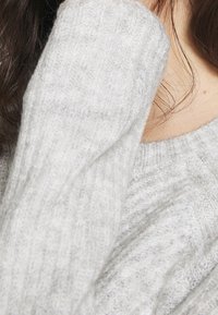 Maglione lavorato a maglia grigio chiaro con una superficie morbida e testurizzata. Dettagli a costine visibili sulle maniche e sul collo. Design accogliente e informale.