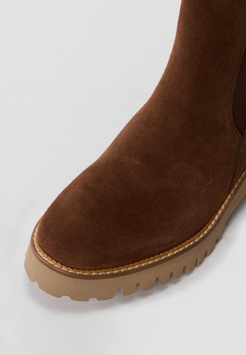 Brązowy zamszowy but z zaokrąglonym noskiem, z teksturowaną cholewką i grubą gumową podeszwą z widocznym bieżnikiem. Krawędź obszyta detalem.