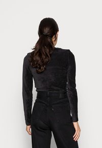 Frau mit langen, dunklen Haaren im Pferdeschwanz, die ein schwarzes langärmliges Oberteil und hochgeschneiderte schwarze Jeans trägt und von einem schlichten Hintergrund abgewandt ist.