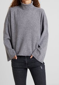 Grijze gebreide coltrui met een relaxed fit, laat vallende schouders en ribbelkraag; gecombineerd met distressed zwarte jeans.
