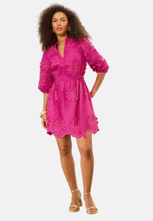 Ženska nosi svetlo roza obleko z vezenimi rožami, s tričetrtinskimi napihnjenimi rokavi in zavezanim pasom, kombinirano z zlatimi sandali in zapestnicami.