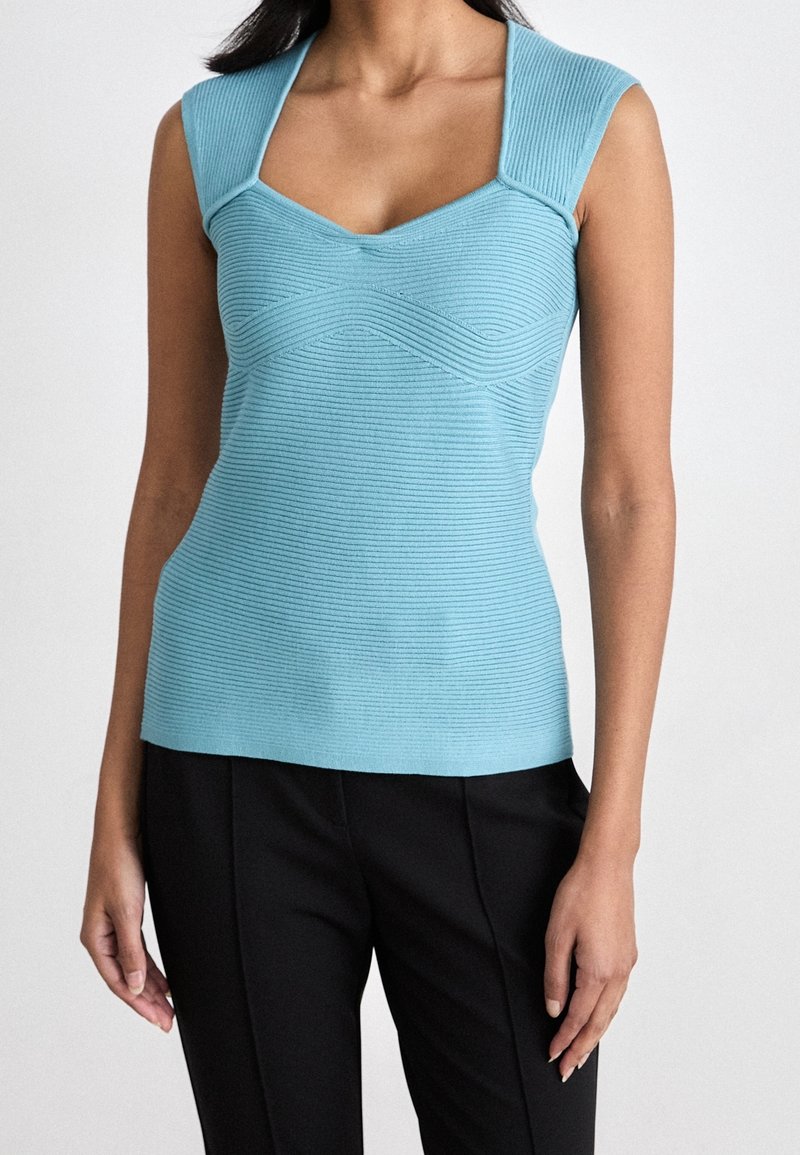 Femme portant un haut en tricot côtelé sans manches bleu clair avec de larges bretelles et un pantalon noir sur un fond clair uni.